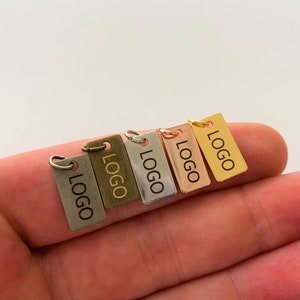 Pode incluir: Cinco pequenas etiquetas metálicas com formato retangular e cantos arredondados. Cada etiqueta tem um furo na parte superior para ser presa a um produto. As etiquetas são de diferentes cores: prata, ouro, bronze, ouro rosa e latão. Cada etiqueta tem a palavra "LOGO" gravada.