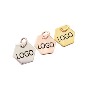 May include: Three hexagonal metal charms in silver, rose gold, and gold. Each charm has a small loop at the top and the word "LOGO" engraved on the front. The charms are displayed against a white background.