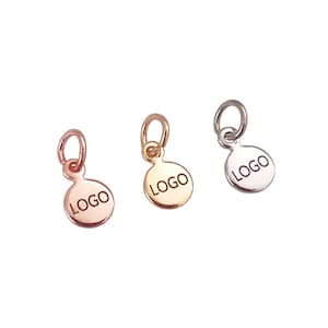 May include: Three small round metal charms with a jump ring. Each charm has the word "LOGO" engraved on the front. The charms are in gold, rose gold, and silver.