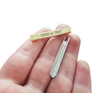 Puede incluir: Una barra de metal dorado con el texto "LOGO or TEXT" grabado en ella. También se muestra una barra de metal plateado con un patrón decorativo.