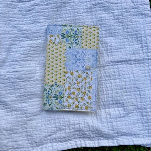 May include: A rectangular fabric book cover featuring a patchwork design of various floral and botanical patterns in shades of yellow, light blue, and white. The cover includes a small white button closure on the right side with a thin elastic loop.