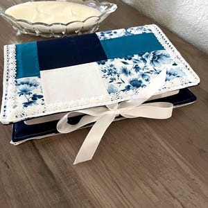May include: A handmade book cover with a patchwork design. The cover features navy blue, teal, and white floral fabric, trimmed with white lace. A white satin ribbon is tied around the cover. The cover is resting on a wooden surface.