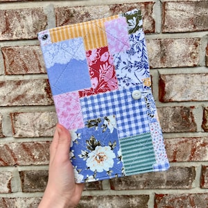 May include: A rectangular fabric book cover featuring a quilted patchwork design with various patterns including blue and white gingham, floral prints in red, pink, and blue, and striped green fabric. A single white button serves as a closure.