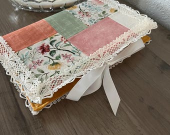 May include: A handmade, quilted book cover with a patchwork design. The cover features squares of floral and solid fabrics in shades of pink, green, and peach, trimmed with lace and tied with a white ribbon. The cover is resting on a wooden surface.