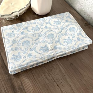May include: A rectangular, quilted fabric case with a floral pattern in light blue on a cream background. The case is closed with a white elastic loop and button. The case is on a wooden surface, with a candle and vase in the background.