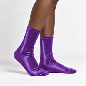 Könnte beinhalten: Ein Paar wadenhohe Socken in einer leuchtenden, einfarbigen violetten Farbe. Der Stoff hat eine glatte, glänzende und leicht reflektierende Oberfläche, die einen satinartigen Look erzeugt. Die Socken haben eine Passform mit Falten.