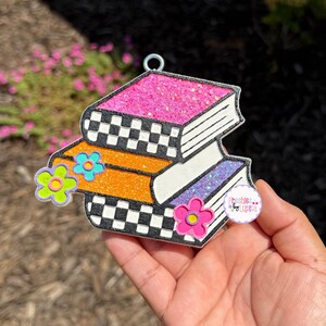 May include: A stack of three glittery books in pink, orange, and purple, adorned with flower accents. The books have a black and white checkered pattern on the edges. A small tag with text is visible.