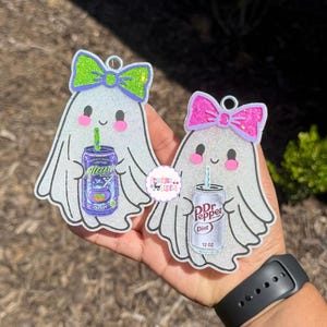 May include: Two glittery white ghost-shaped ornaments with hanging loops. The left ghost wears a lime green bow and holds a purple Alani Nu energy drink. The right ghost wears a hot pink bow and holds a silver Diet Dr Pepper can.