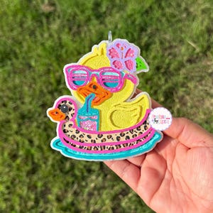 May include: A yellow duck-shaped air freshener with pink sunglasses, a flower, and a drink. The duck is in a leopard print floatie with a turquoise border. The air freshener is held in a hand.