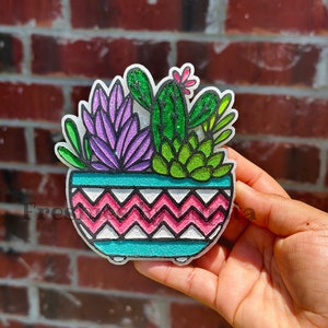 May include: A colorful ceramic planter with a pink and white chevron pattern. The planter is filled with green and purple succulents and a cactus with a pink flower.
