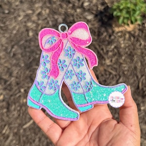May include: A decorative ornament featuring a pair of cowboy boots and a bow. The boots are turquoise with glitter, purple and white floral details, and a pink outline. The bow is pink with glitter.