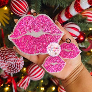 May include: Two pink glitter lips ornaments with a white background. The ornaments are hanging from a Christmas tree with candy cane ornaments and red and white striped ornaments. The ornaments are made by "Froshies by Lupita".