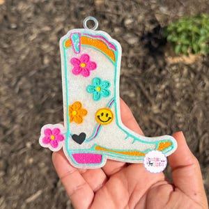 May include: A white cowboy boot-shaped ornament with a turquoise outline, adorned with colorful flowers, a smiley face, and a black heart. The boot has a glittery orange and turquoise top and a pink sole. A metal ring is attached for hanging.