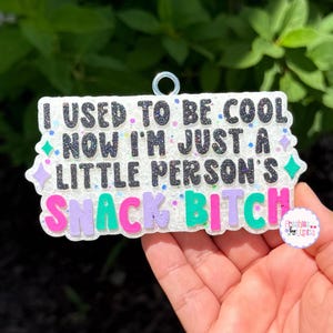 May include: A rectangular white glittery air freshener with a silver hanging loop. The text reads: I USED TO BE COOL NOW I'M JUST A LITTLE PERSON'S SNACK BITCH. The words are in black, purple, and teal with small star accents and a small logo sticker.