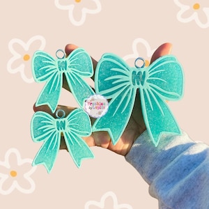 May include: Three turquoise blue glitter bow charms with silver jump rings. The bows are different sizes and are held in a hand.