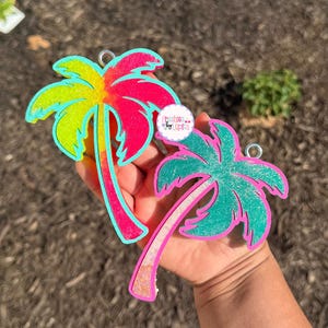 May include: Two colorful palm tree-shaped ornaments. One is a gradient of yellow, orange, and pink with a teal outline. The other is teal with a pink outline. Both have a silver ring for hanging. The text "Freshies... by Lupita" is visible.