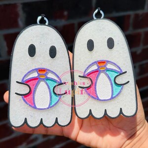 May include: Two white ghost-shaped ornaments with black outlines, each holding a pink, blue, and white beach ball. The ornaments are covered in glitter.