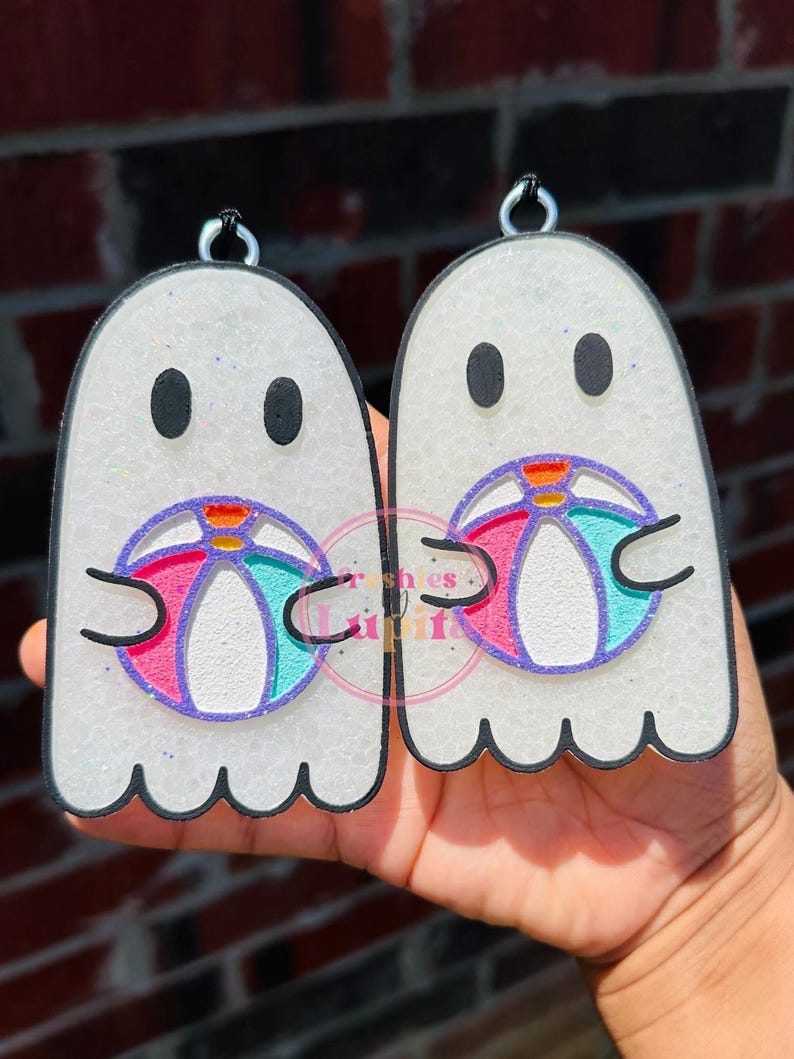 May include: Two white ghost-shaped ornaments with black outlines, each holding a pink, blue, and white beach ball. The ornaments are covered in glitter.