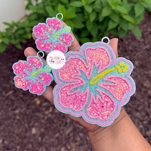 May include: Three glittery hibiscus flower ornaments in varying sizes. The flowers feature pink petals with glitter, outlined in a light blue and pink border. The center of the flowers are yellow. The text "Freshies by Lupita" is visible.