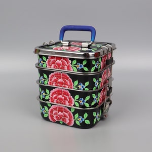 Op de afbeelding: Een zwarte metalen lunchbox met een blauwe handgreep en een bloemenontwerp. De lunchbox heeft vier lagen en is versierd met roze en groene bloemen.