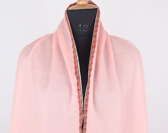 Puede incluir: Un chal rosa pálido con un delicado borde bordado en tonos rojos, verdes y crema. El chal está drapeado sobre un maniquí y presenta motivos inspirados en el paisley.