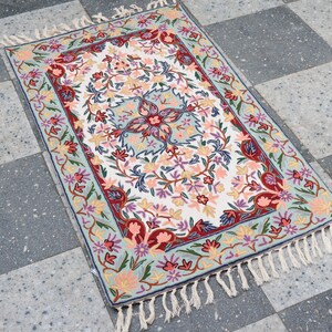 Puede incluir: Alfombra rectangular con centro blanco y diseño floral en rojo, rosa y azul. La alfombra tiene un borde azul claro y flecos blancos. La alfombra está colocada sobre un suelo de baldosas a cuadros grises y blancos.