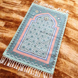 May include: A turquoise prayer rug with a geometric pattern and a pink and blue border. The rug has a white fringe around the edges.