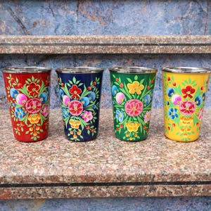 Puede incluir: Cuatro vasos de acero inoxidable pintados a mano en rojo, azul marino, verde y amarillo. Cada vaso presenta un diseño floral con flores rosas, azules y amarillas. Los vasos tienen un borde plateado y se exhiben sobre una superficie de piedra.