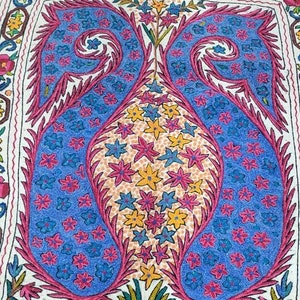 May include: A white fabric rug with a colorful embroidered design. The design features a vase with flowers and leaves, all in shades of pink, blue, yellow, and green.