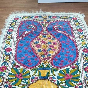 May include: A white embroidered rug with a floral design featuring a vase with two peacocks. The rug has a border of flowers and leaves in shades of pink, yellow, blue, and green.