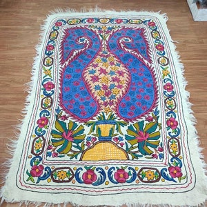 May include: A white rug with a colorful floral design. The rug has a fringed edge and features a central motif of two blue paisley shapes with yellow, pink, and green flowers.