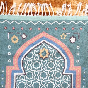 May include: A teal blue prayer rug with a geometric pattern in the center. The rug has a pink, blue, and white border with a fringe on the top.