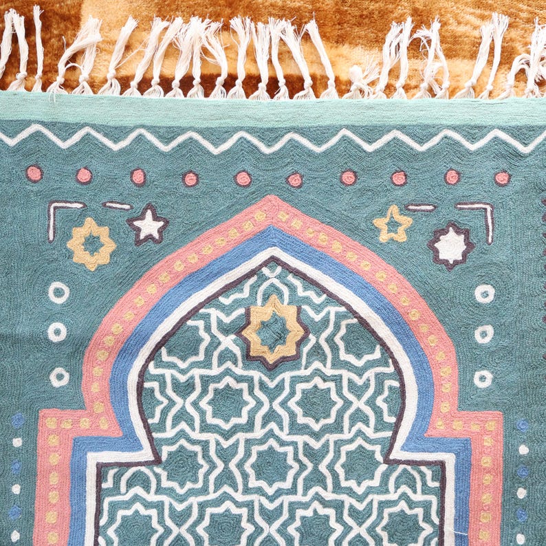 May include: A teal blue prayer rug with a geometric pattern in the center. The rug has a pink, blue, and white border with a fringe on the top.