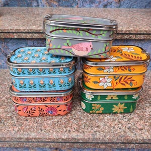 May include: A stack of colorful metal lunchboxes with floral and bird designs. The lunchboxes are painted in various colors, including gray, blue, orange, yellow, and green. Each box has a stainless steel handle and trim.
