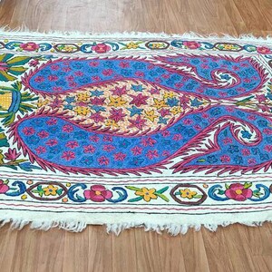 May include: A white rug with a colorful embroidered design featuring a paisley pattern in shades of blue, pink, yellow, and green. The rug has a fringed edge.