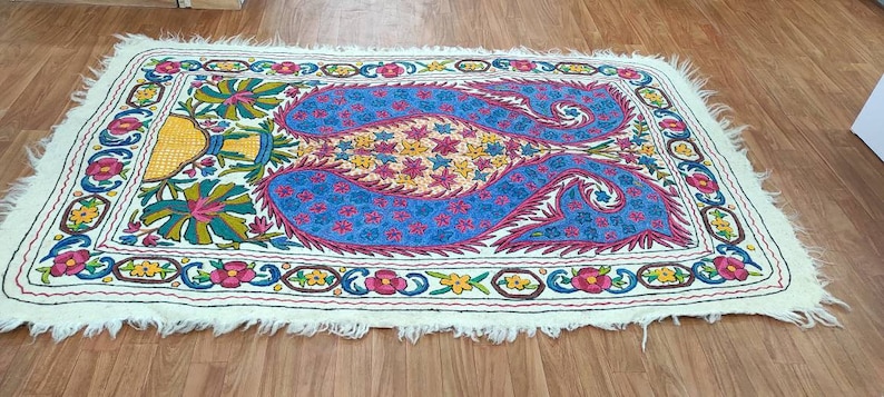 May include: A white rug with a colorful embroidered design featuring a paisley pattern in shades of blue, pink, yellow, and green. The rug has a fringed edge.