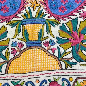 May include: A white fabric square with colorful embroidery depicting a vase with flowers and leaves. The embroidery is done in shades of pink, blue, green, yellow, and brown.