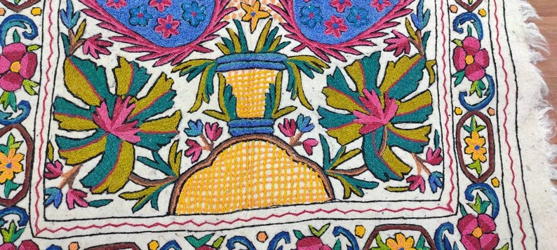 May include: A white fabric square with colorful embroidery depicting a vase with flowers and leaves. The embroidery is done in shades of pink, blue, green, yellow, and brown.