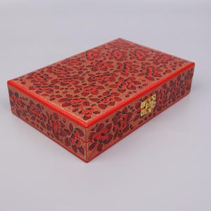 May include: A red wooden box with a floral design and a gold latch. The box is closed and sitting on a white surface.