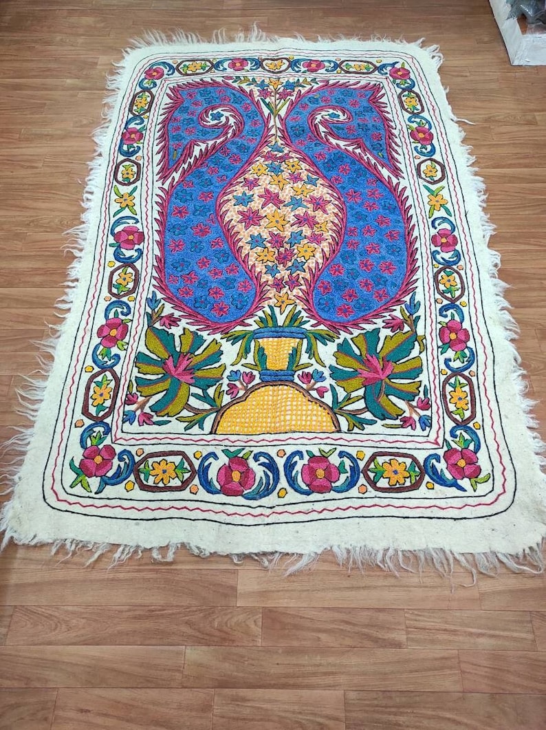 May include: A white rug with a colorful floral design. The rug has a fringed edge and features a central motif of two blue paisley shapes with yellow, pink, and green flowers.