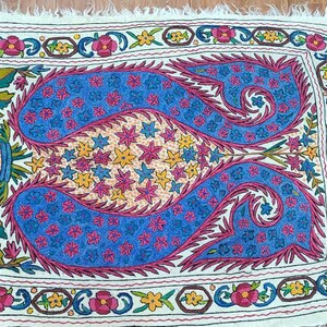 May include: A white fabric rug with a colorful embroidered design. The design features two blue paisley shapes with pink, yellow, and white floral accents. The rug has a fringed border.