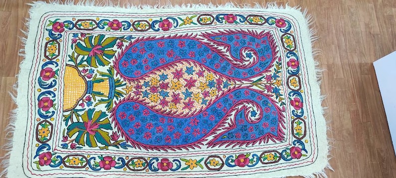 May include: A white fabric rug with a colorful embroidered design. The design features two blue paisley shapes with pink, yellow, and white floral accents. The rug has a fringed border.