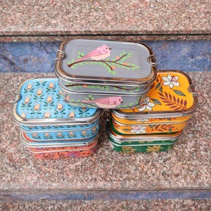 May include: A stack of colorful stainless steel lunch boxes. The lids feature various designs, including a pink bird on a branch, floral patterns in blue, orange, yellow, and green. Each box has a silver clasp.