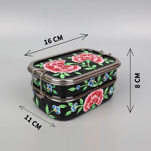 May include: A black, two-tiered lunchbox with a floral design. The lunchbox is 16 cm long, 11 cm wide, and 8 cm tall. It features a stainless steel trim and clasp, and is decorated with red and blue flowers.
