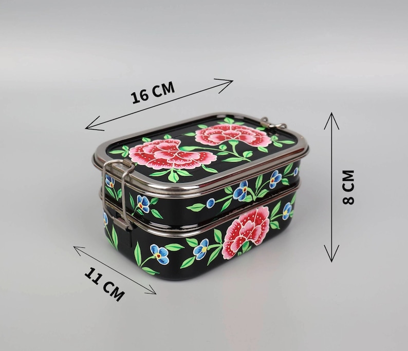 May include: A black, two-tiered lunchbox with a floral design. The lunchbox is 16 cm long, 11 cm wide, and 8 cm tall. It features a stainless steel trim and clasp, and is decorated with red and blue flowers.