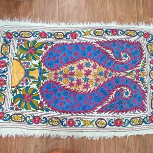 May include: A white rug with a colorful embroidered design featuring paisley patterns, flowers, and stars. The rug has a fringed edge.