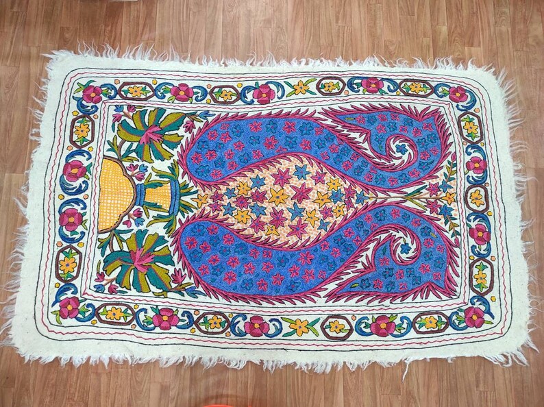 May include: A white rug with a colorful embroidered design featuring paisley patterns, flowers, and stars. The rug has a fringed edge.