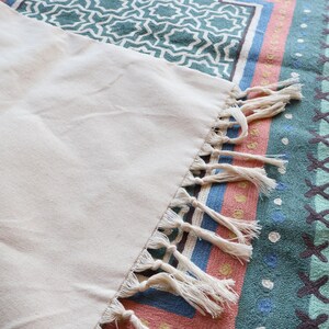 May include: A white cotton throw blanket with fringe trim. The blanket is laying on a colorful patterned rug.