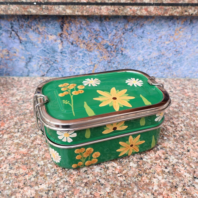 May include: A two-tiered, green metal lunchbox with a stainless steel clasp and trim. The lunchbox is decorated with hand-painted yellow and white floral designs. The lunchbox is sitting on a speckled, brown surface.