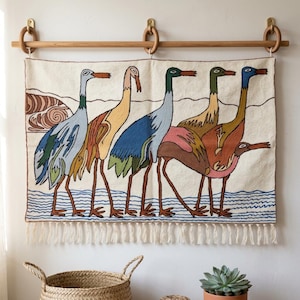 May include: A rectangular wall hanging featuring a woven design of five stylised birds in shades of blue, yellow, green, and brown standing in blue water. The textile has a cream background, a brown border, and a fringe along the bottom edge.